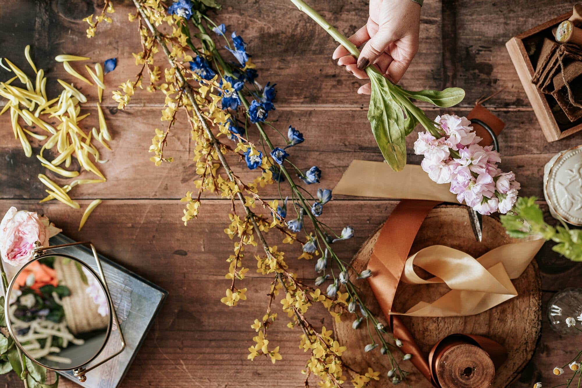 Mains tenant une fleur rose parmi branches jaunes et bleues, rubans et ciseaux sur une table