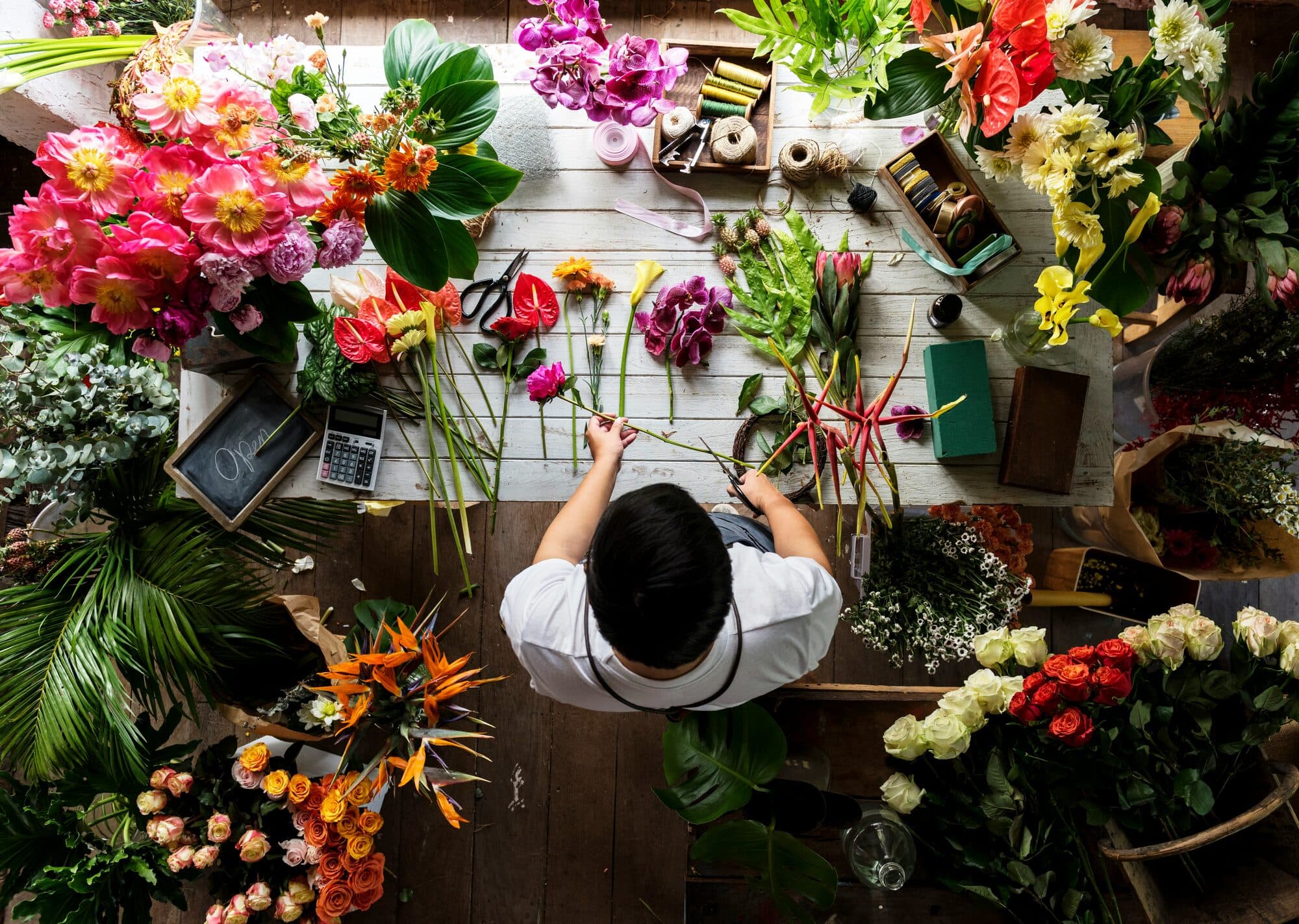 Fleuriste coupant des tiges au milieu de pivoines, roses et feuillages sur un établi