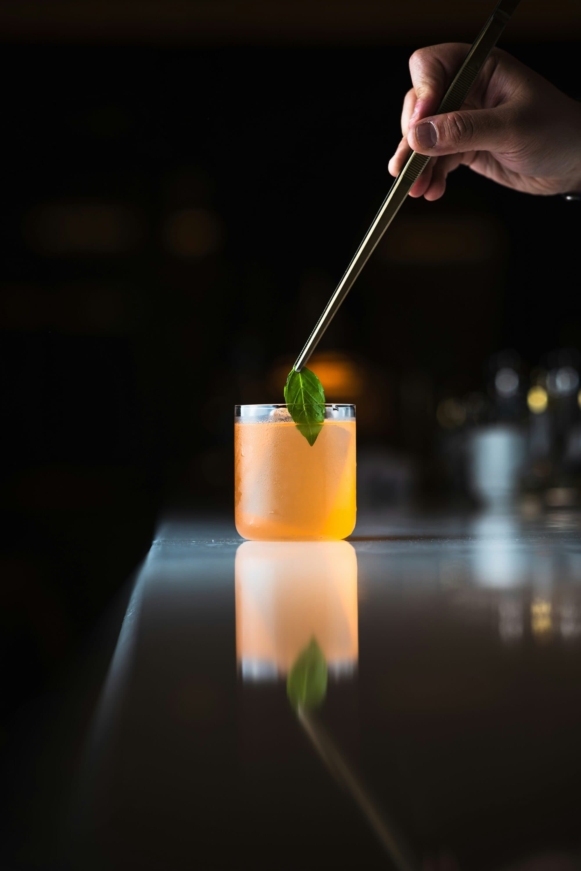 Main déposant une feuille verte avec une pince sur un cocktail orangé au comptoir